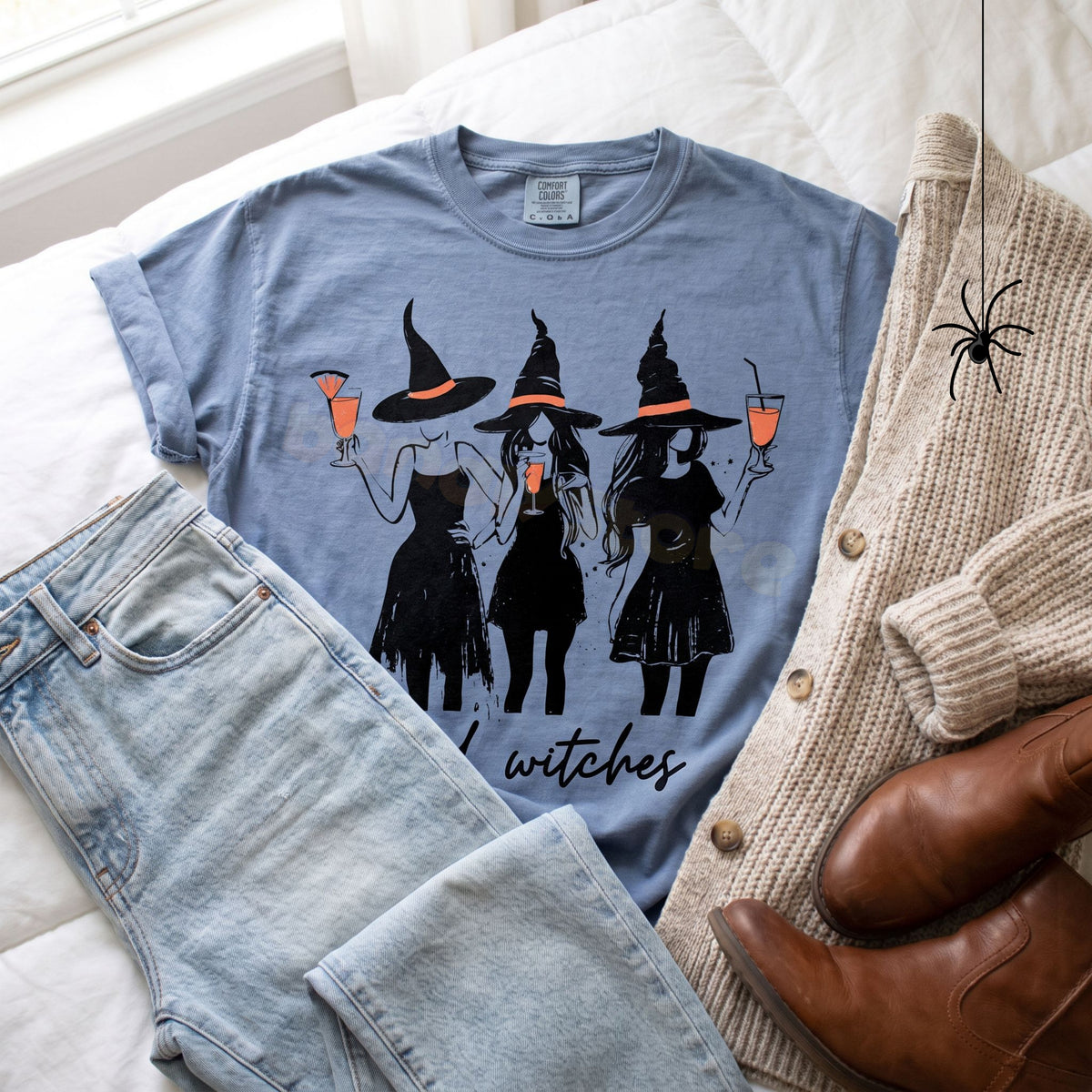 Boujee Bad Witches T-Shirt in Blue Jean flat-laid on white bedding with jeans, beige cardigan, brown boots and a spider.