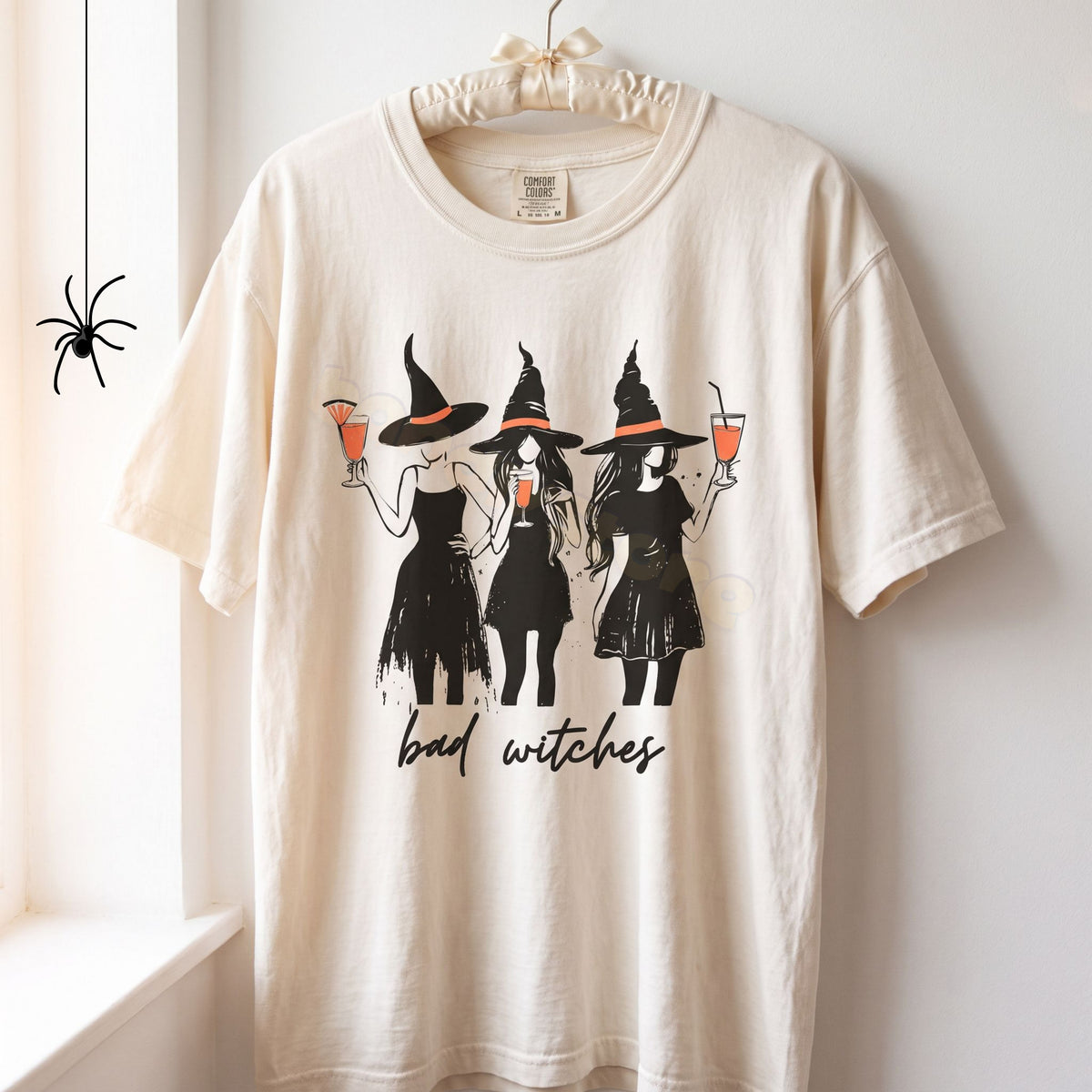 Boujee Bad Witches T-Shirt in ivory hangs on a wall hook by a window with a small spider graphic nearby.