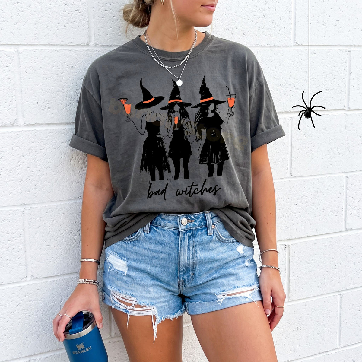 Boujee Bad Witches T-Shirt in pepper worn front-facing against a white brick wall holding a blue tumbler near a spider.