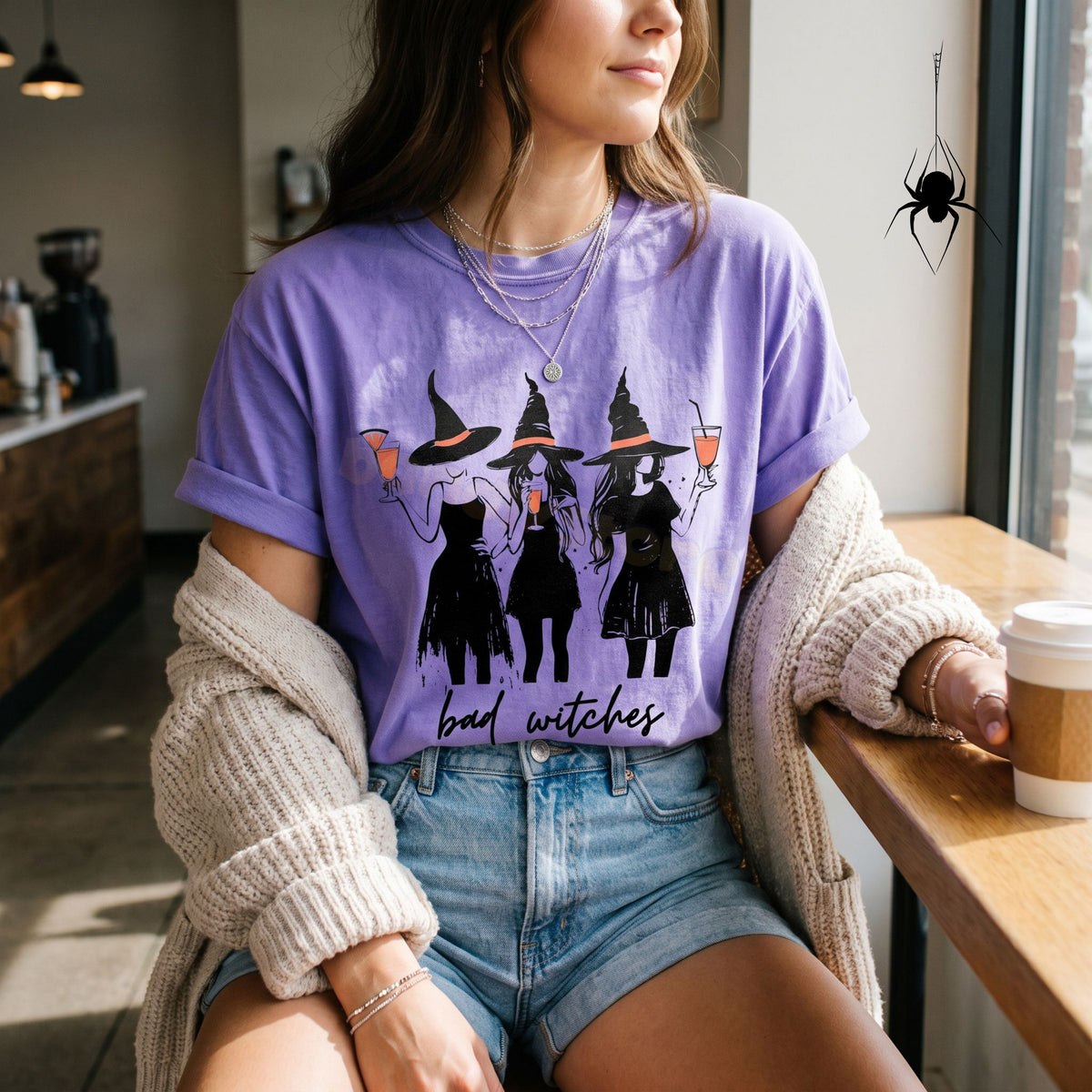 Boujee Bad Witches T-Shirt in violet worn front-facing by a person seated in a cafe by a window with coffee cup.