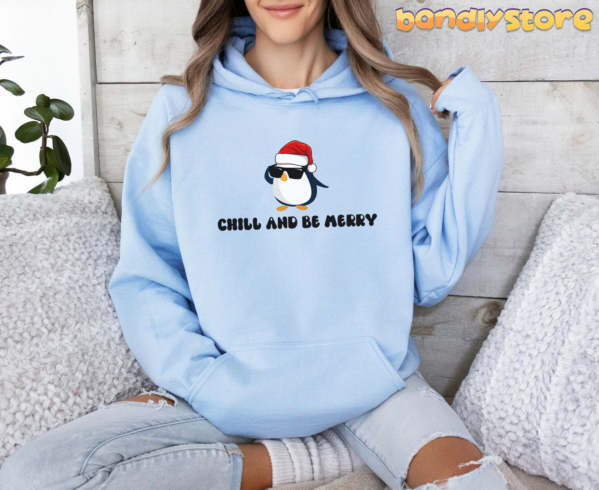Chill And Be Merry Penguin Hoodie in light blue, worn front-facing by a seated person amid pillows and a plant.