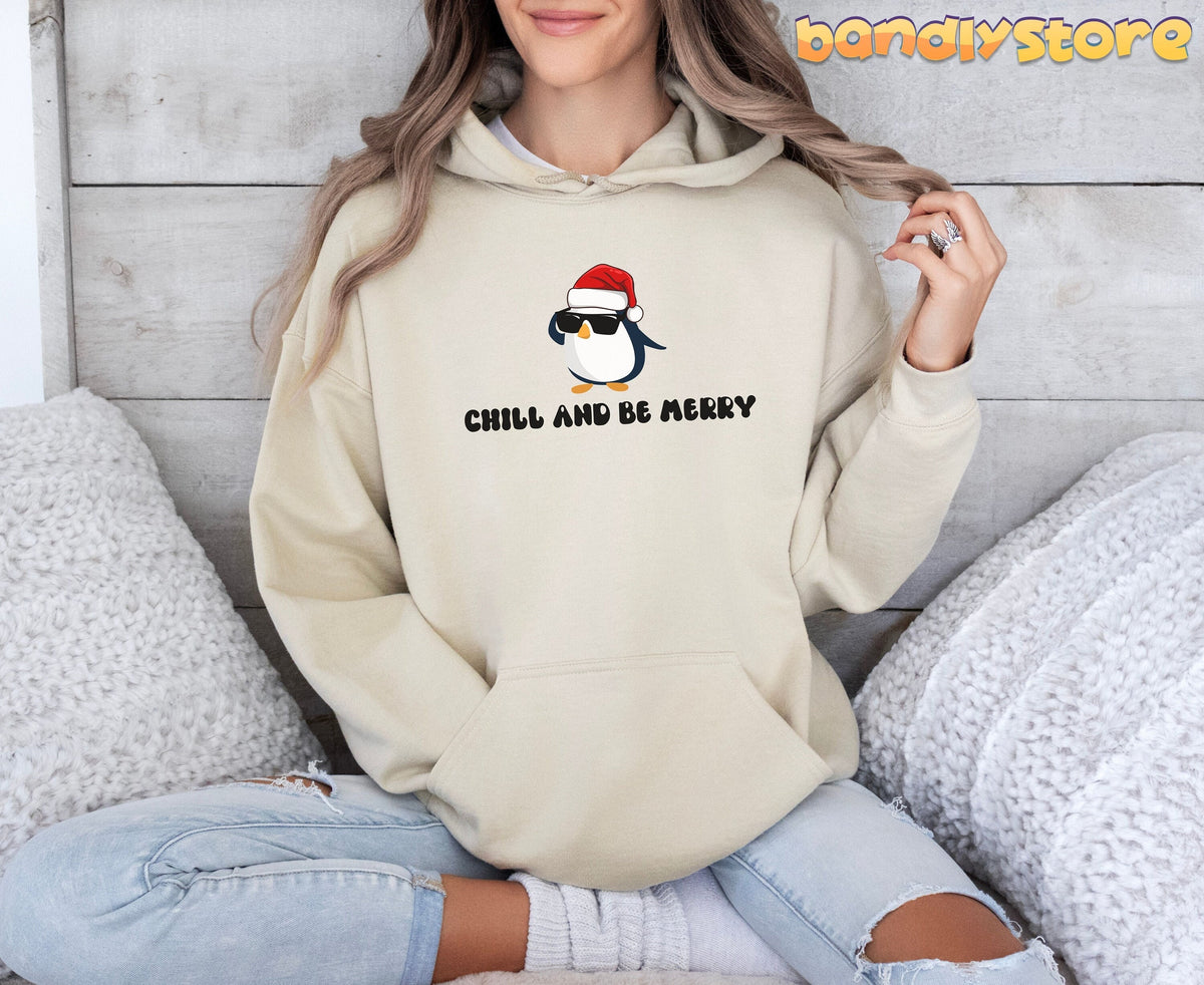 Chill And Be Merry Penguin Hoodie in sand on a person sitting cross-legged amid white fluffy pillows by a wood wall.
