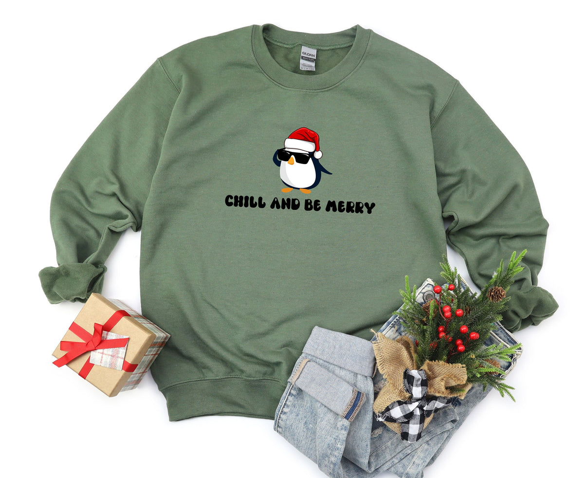 Chill And Be Merry Penguin Sweatshirt in military green, flat-lay with gift box, jeans and greenery.