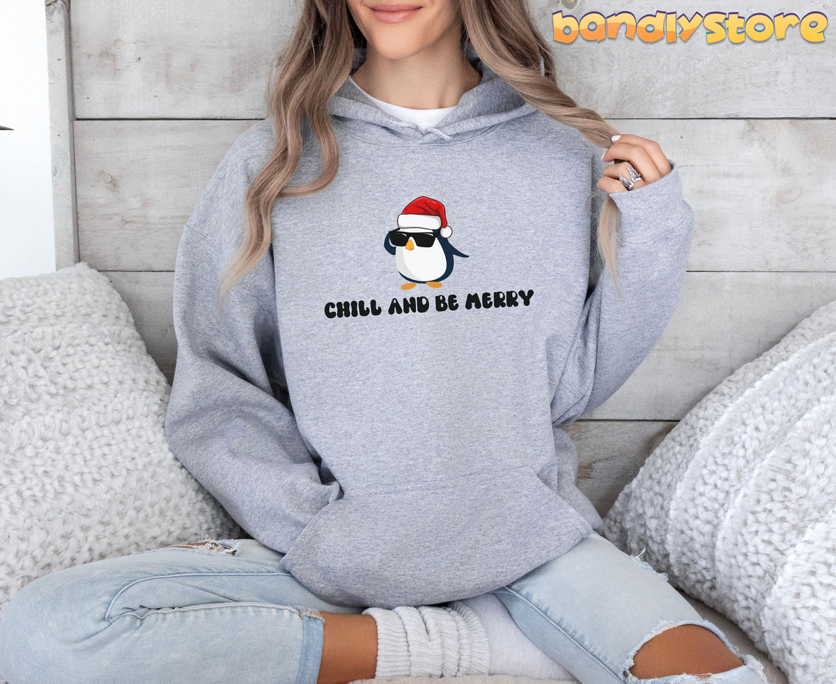 Chill And Be Merry Penguin Hoodie in Sport Grey on a seated person against wood wall with white pillows.