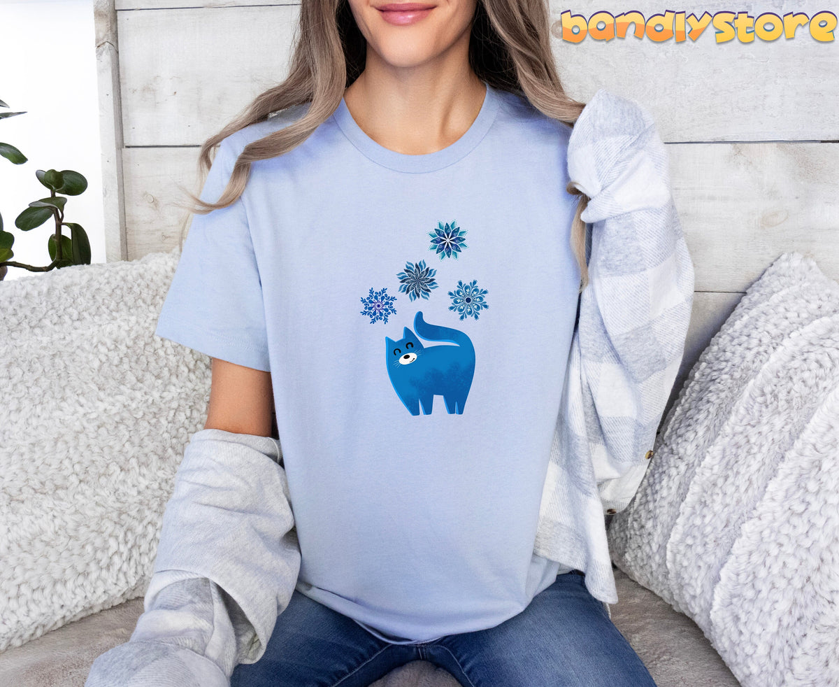 Christmas Cat and Snowflakes T-Shirt in light blue on a person seated on a couch with pillows and a plant.