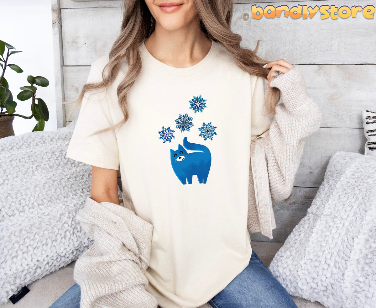 Christmas Cat and Snowflakes T-Shirt in natural on a woman seated amid fluffy pillows against a wooden wall.