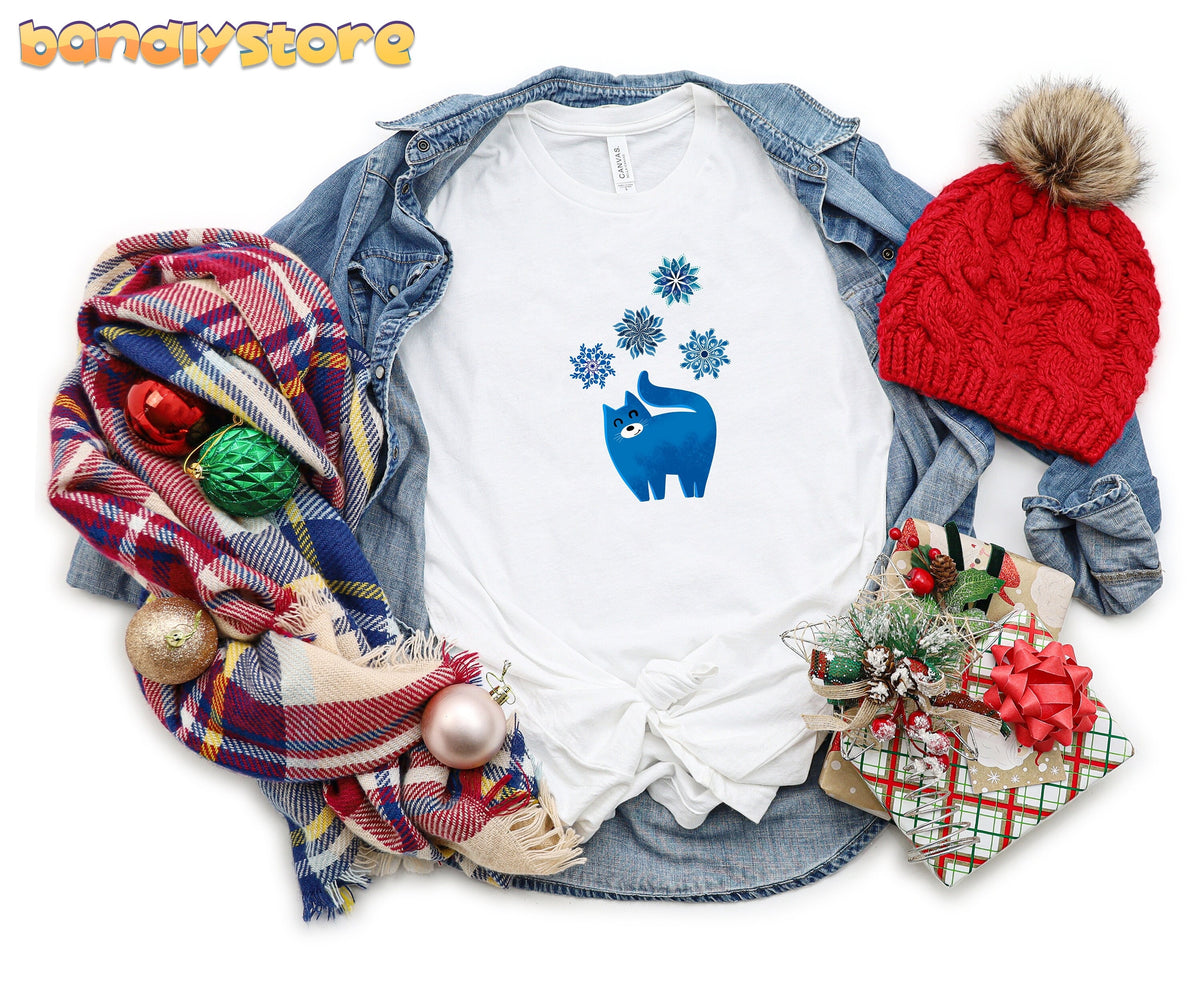 Christmas Cat and Snowflakes T-Shirt white flat lay with denim jacket, plaid scarf, red beanie, ornaments and gifts