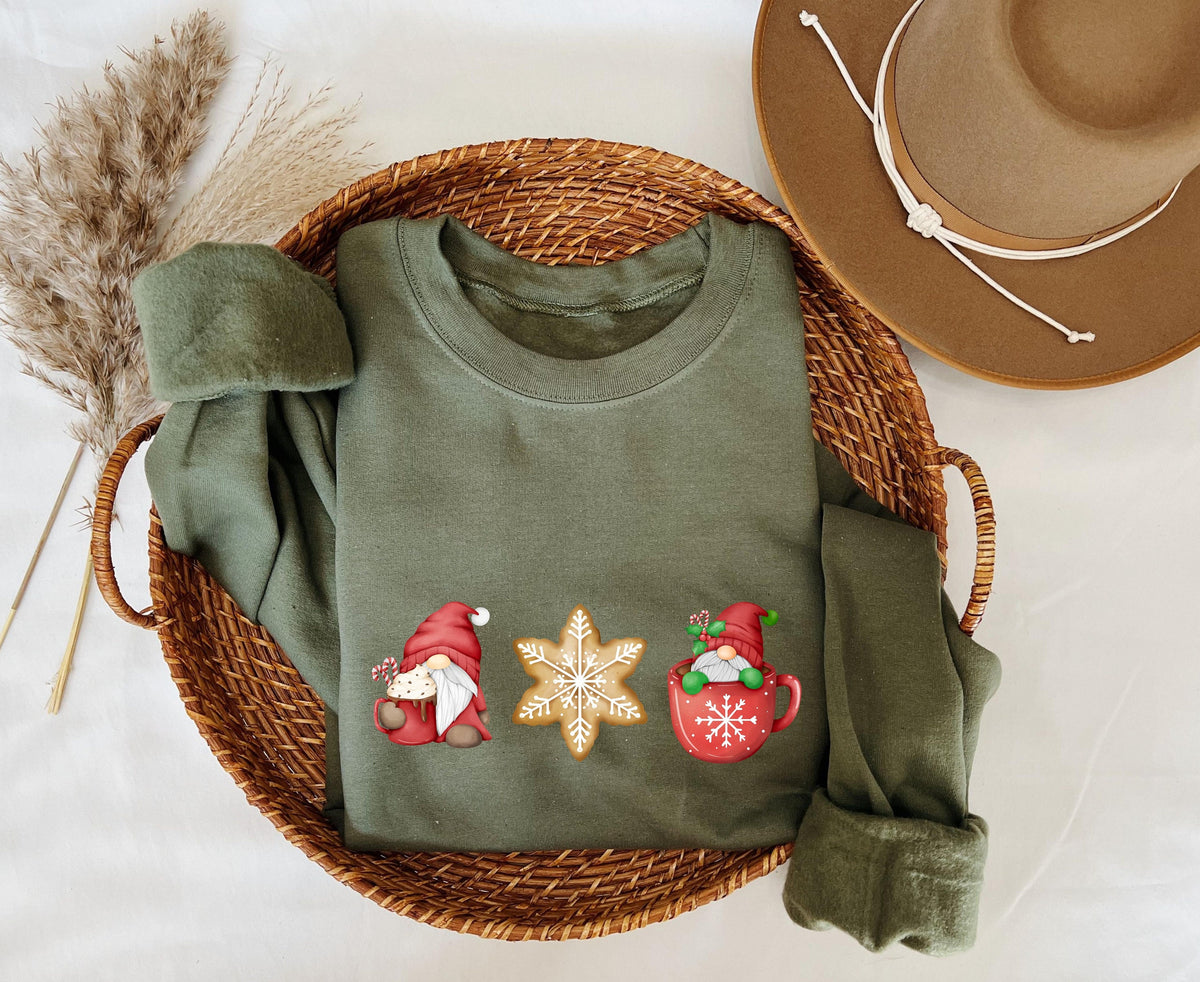 Christmas Gnomes Gingerbread Sweatshirt military green folded in a wicker basket beside dried grass and a hat, top-down view.