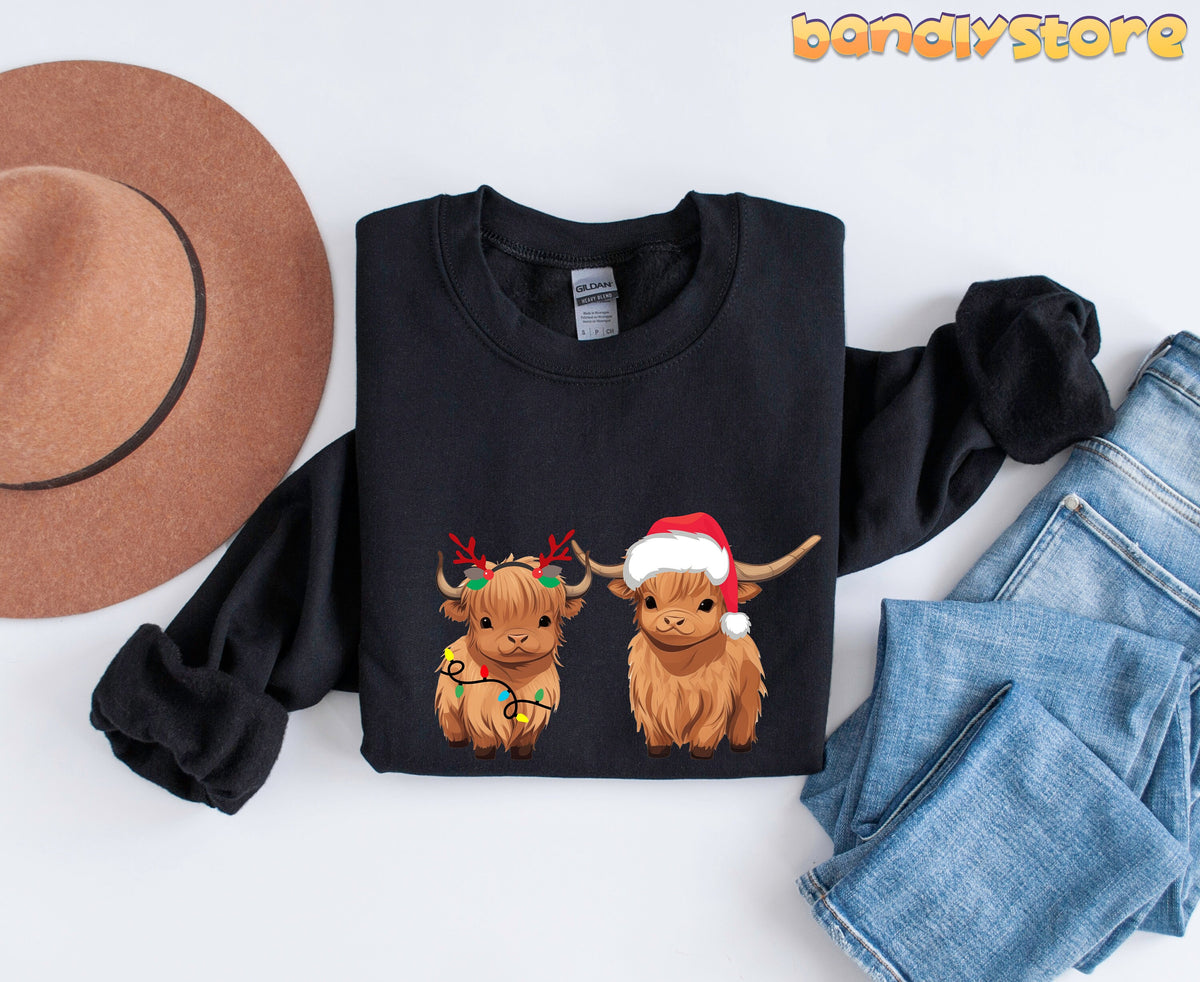Christmas Highland Cow Sweatshirt in black folded flat from above beside a brown hat and blue jeans.