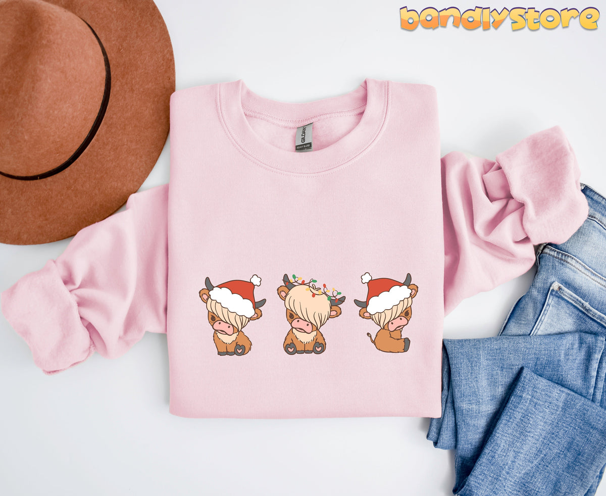 Christmas Highland Cow Trio Sweatshirt in light pink folded flat beside a brown hat and blue jeans.