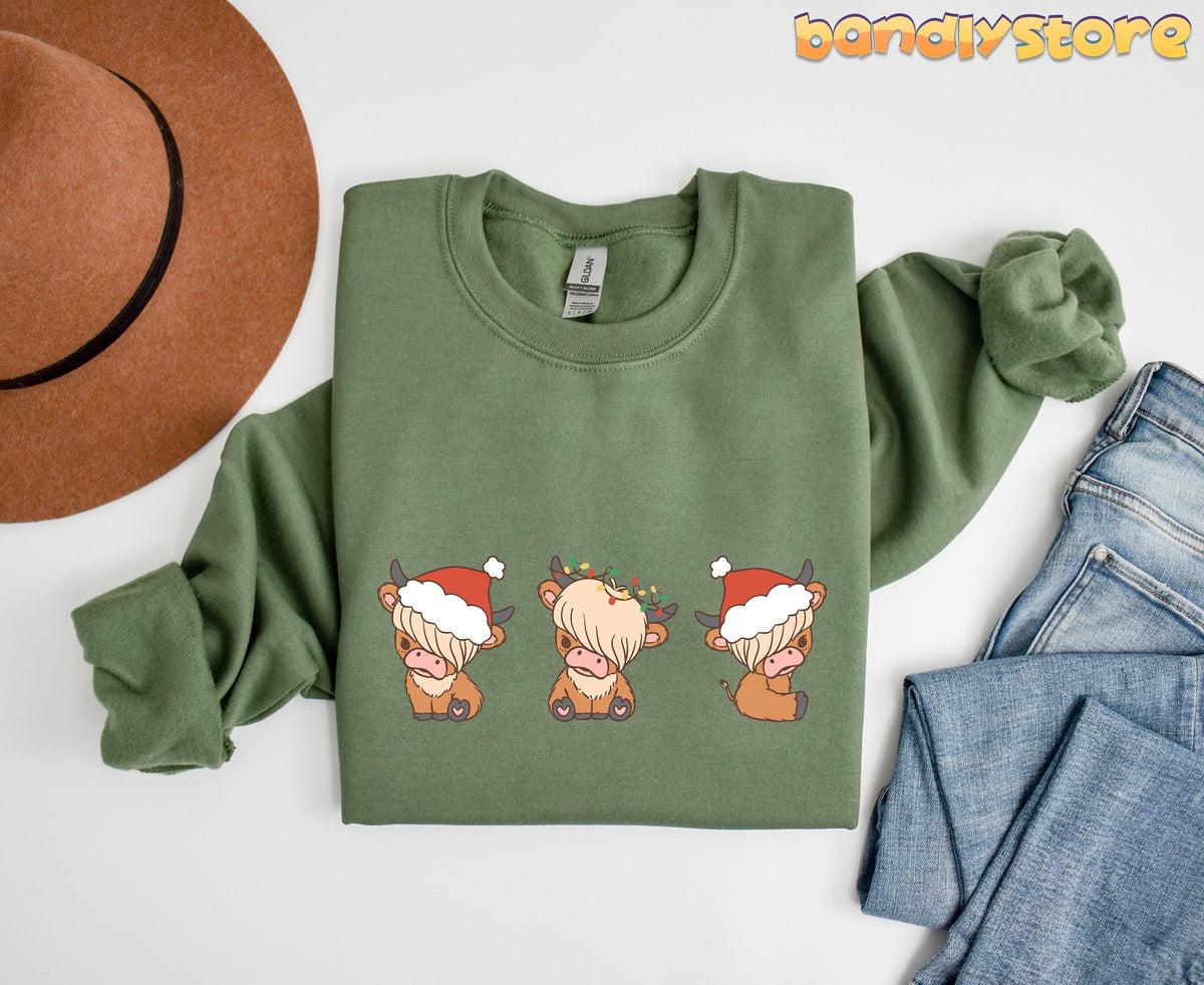 Christmas Highland Cow Trio Sweatshirt in military green shown flat-laid from above beside a brown hat and blue jeans.