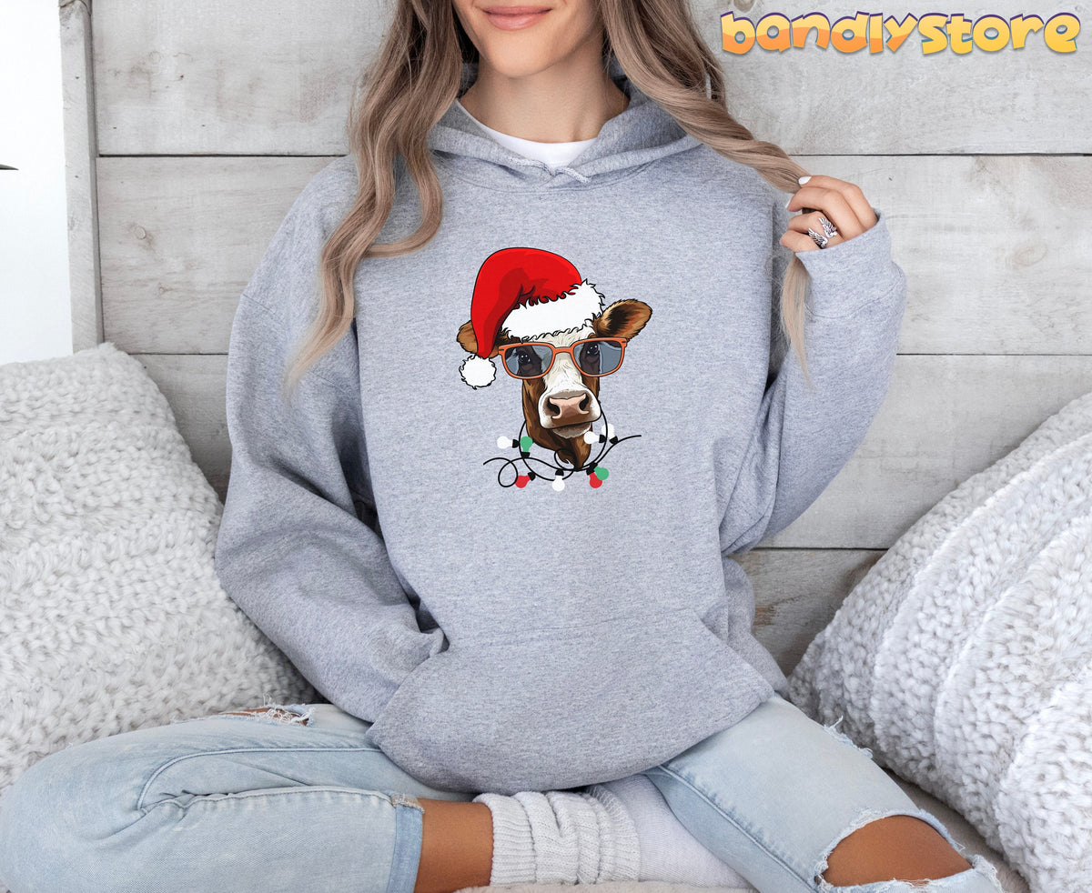 Christmas Highland Cow Hoodie in Sport Grey on a seated person facing forward by white pillows and wood wall.