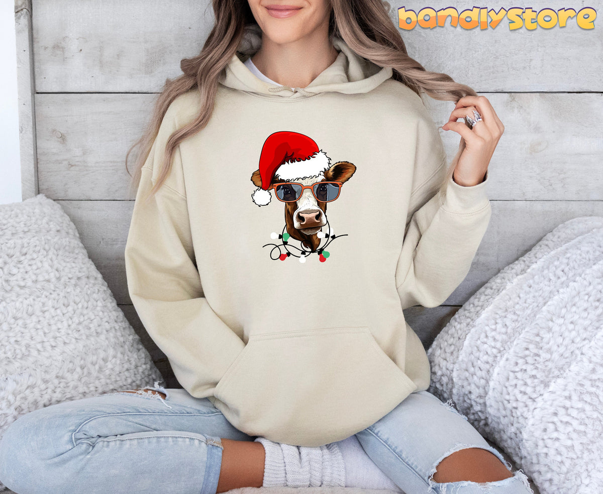 Christmas Highland Cow Hoodie in sand worn by a seated person against a wooden wall with white pillows.
