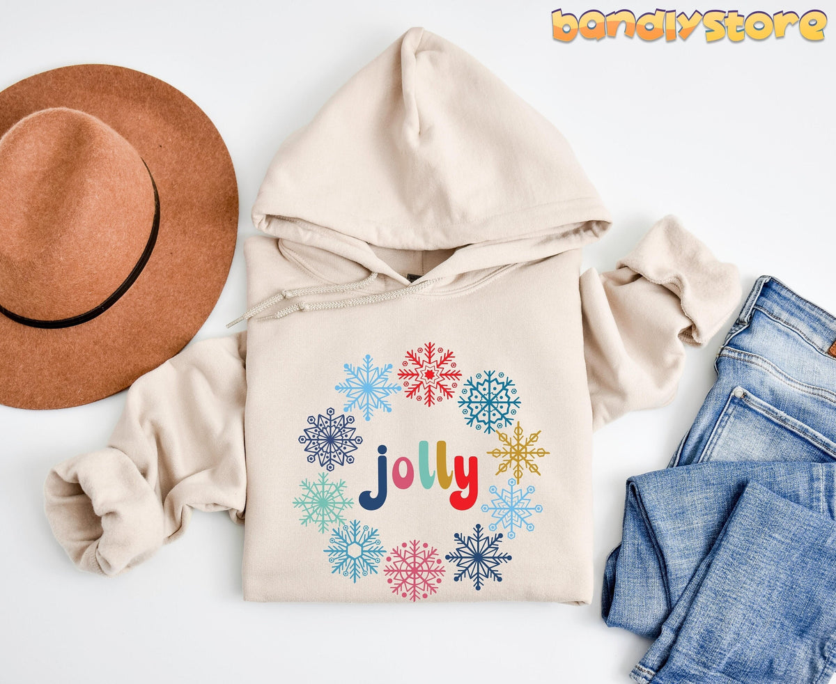 Christmas Snowflake Jolly Hoodie in sand laid flat beside a brown hat and blue jeans.
