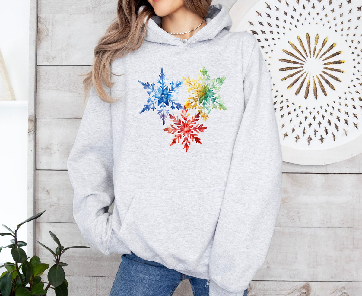 Colorful Watercolor Snowflakes Hoodie in ash, front view on a person amid light wood walls, plants and round wall art.