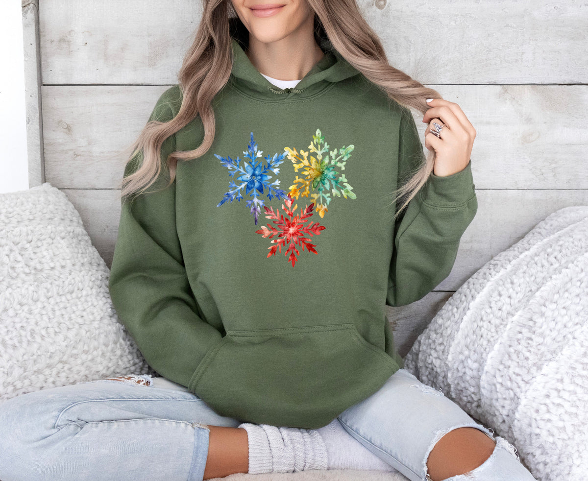Colorful Watercolor Snowflakes Hoodie in military green on a seated person against wood wall with white pillows.