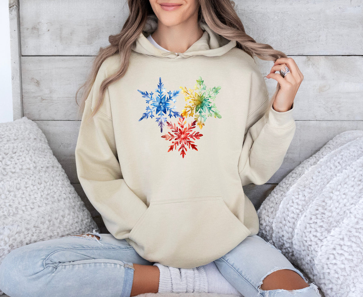 Colorful Watercolor Snowflakes Hoodie in sand, worn seated against a white wood wall with fluffy pillows.
