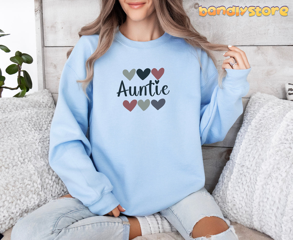 Auntie Sweatshirt in light blue front view on a woman seated among white pillows against wood wall with plant.