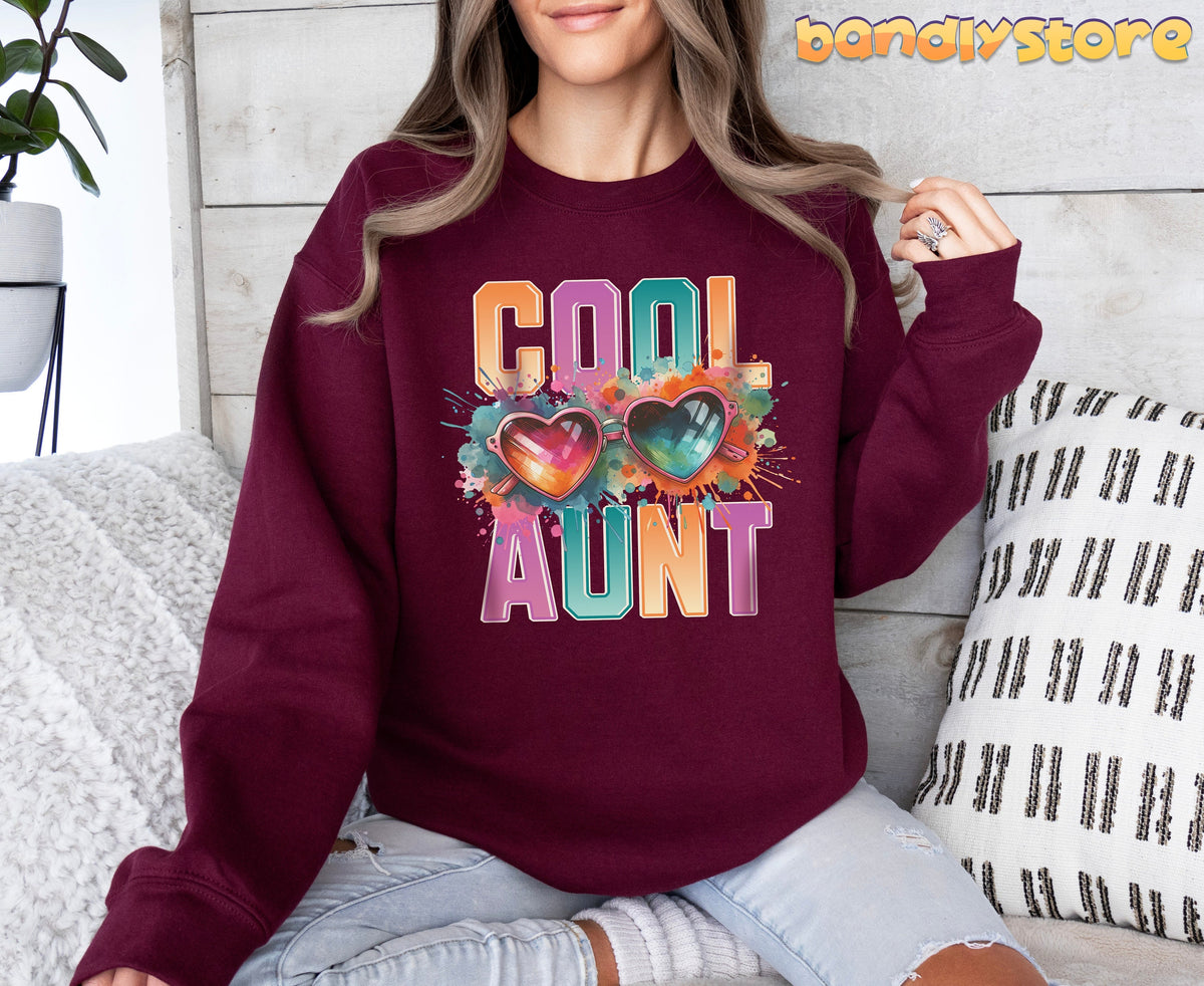 Cool Aunt Sweatshirt in maroon, modeled indoors on a sofa with pillows, plant, and wood wall behind.