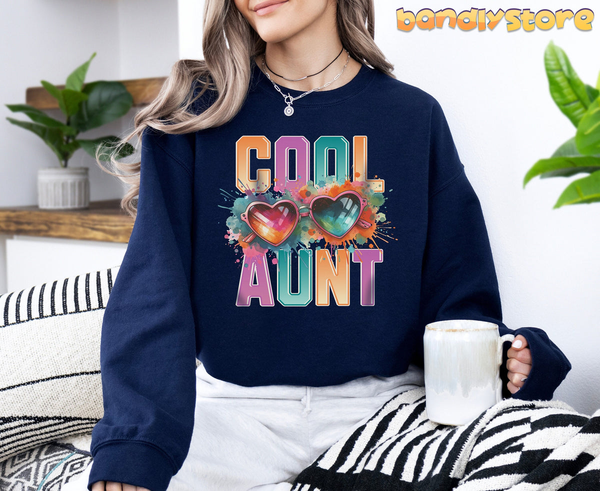 Cool Aunt Sweatshirt in navy on a seated woman holding a mug beside plants and a wooden shelf.