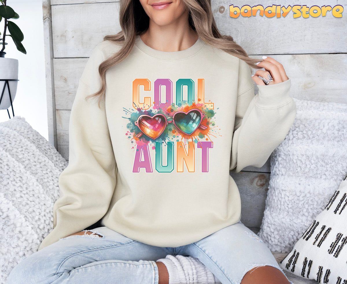 Cool Aunt Sweatshirt in sand on a woman seated amid sofa pillows against a light wood wall.
