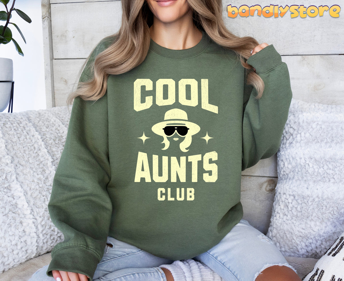 Cool Aunts Club Sweatshirt in military green worn front-on by a person on a sofa with pillows, wood wall and plant.