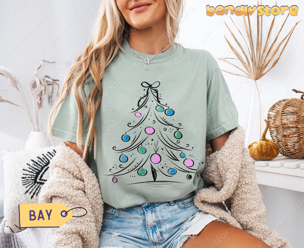 Coquette Christmas Tree T-Shirt in bay on seated model facing camera amid dried plants, pillow and pumpkins.