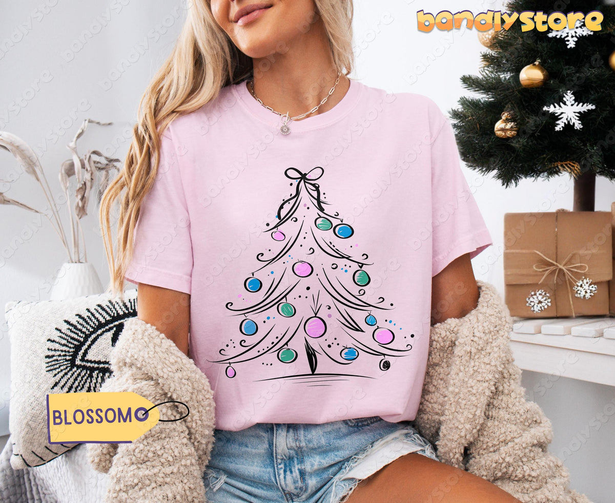 Coquette Christmas Tree T-Shirt in Blossom on a model seated by a Christmas tree with gifts nearby.