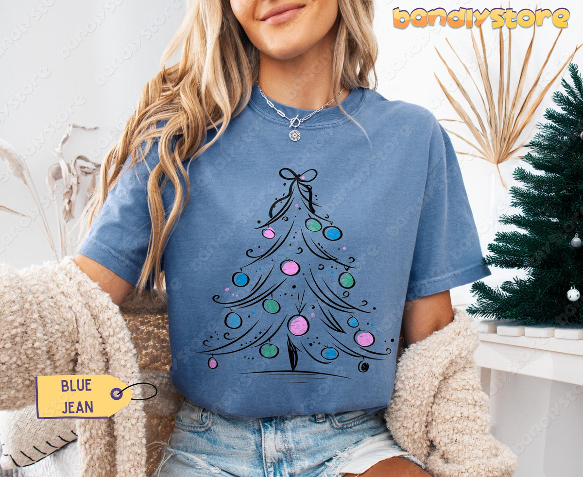 Coquette Christmas Tree T-Shirt in Blue Jean front view on a woman seated by a Christmas tree and dried plants.