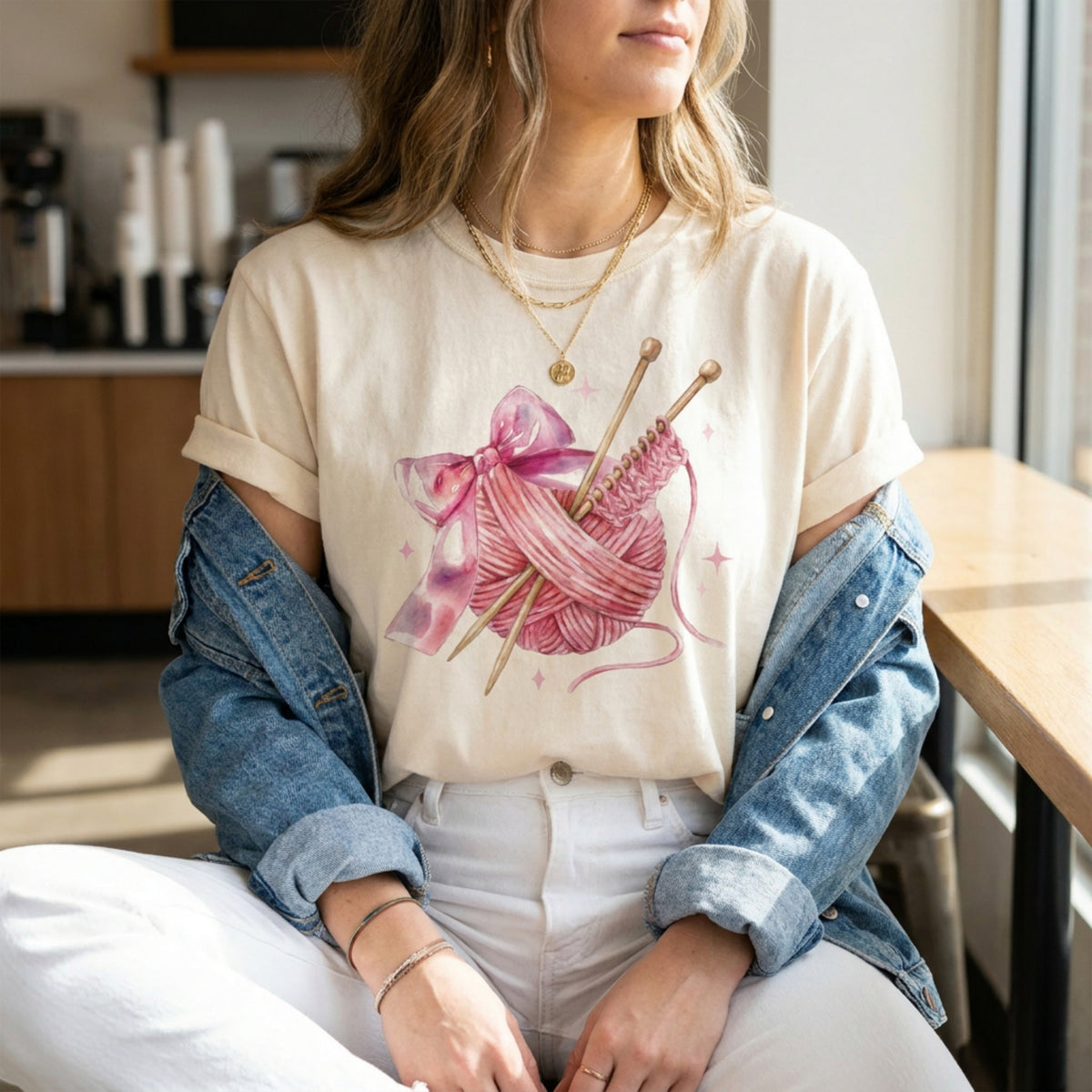 Cute Coquette Knitting T-Shirt in ivory on a seated woman in a cafe with denim jacket and white pants.