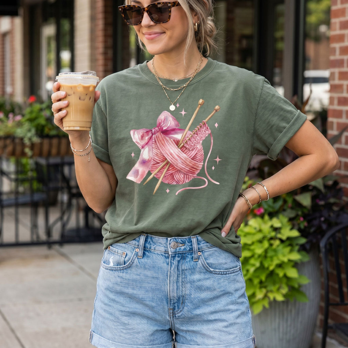 Cute Coquette Knitting T-Shirt in moss on woman holding iced coffee outdoors by plants and brick.