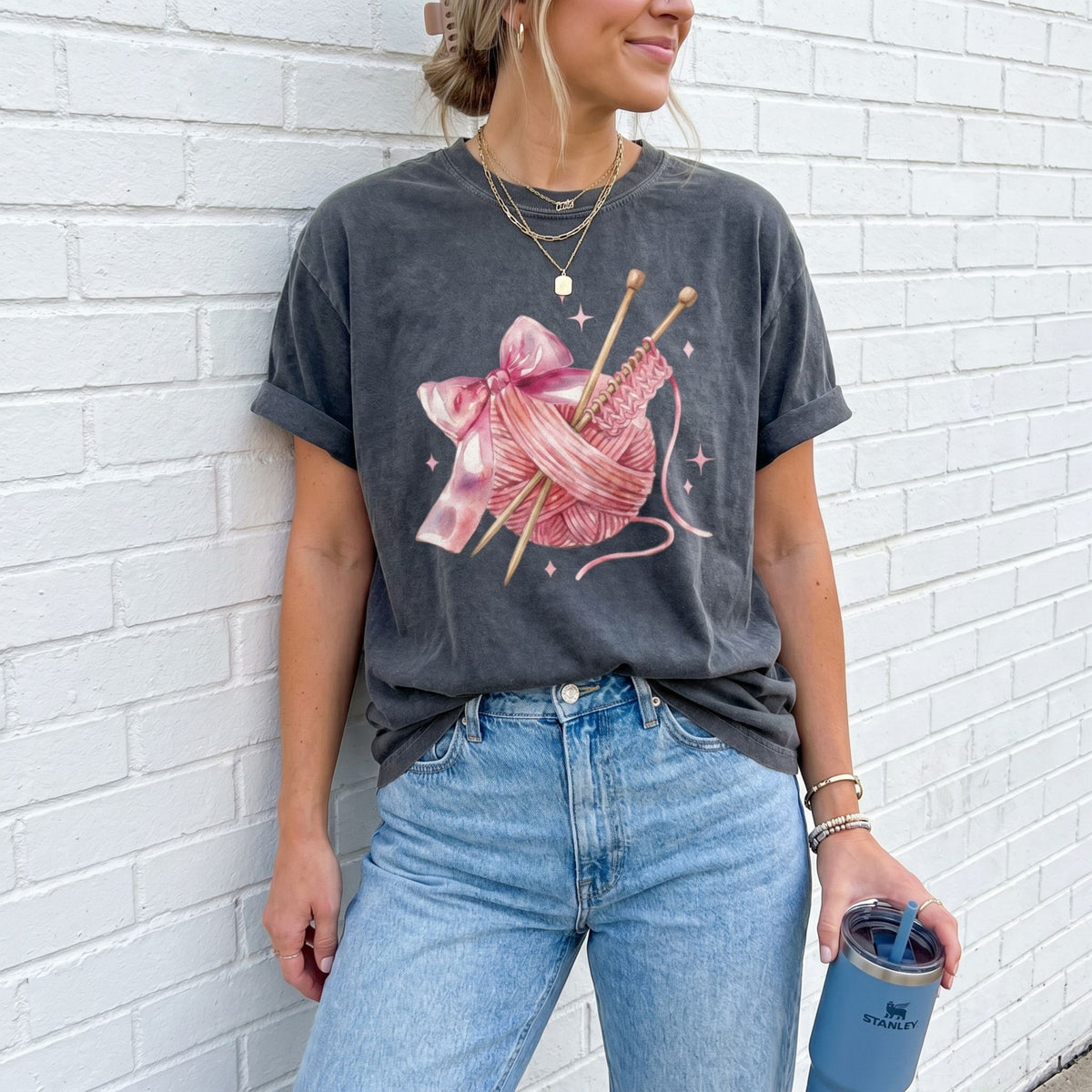 Cute Coquette Knitting T-Shirt in pepper on woman against white brick wall holding tumbler.