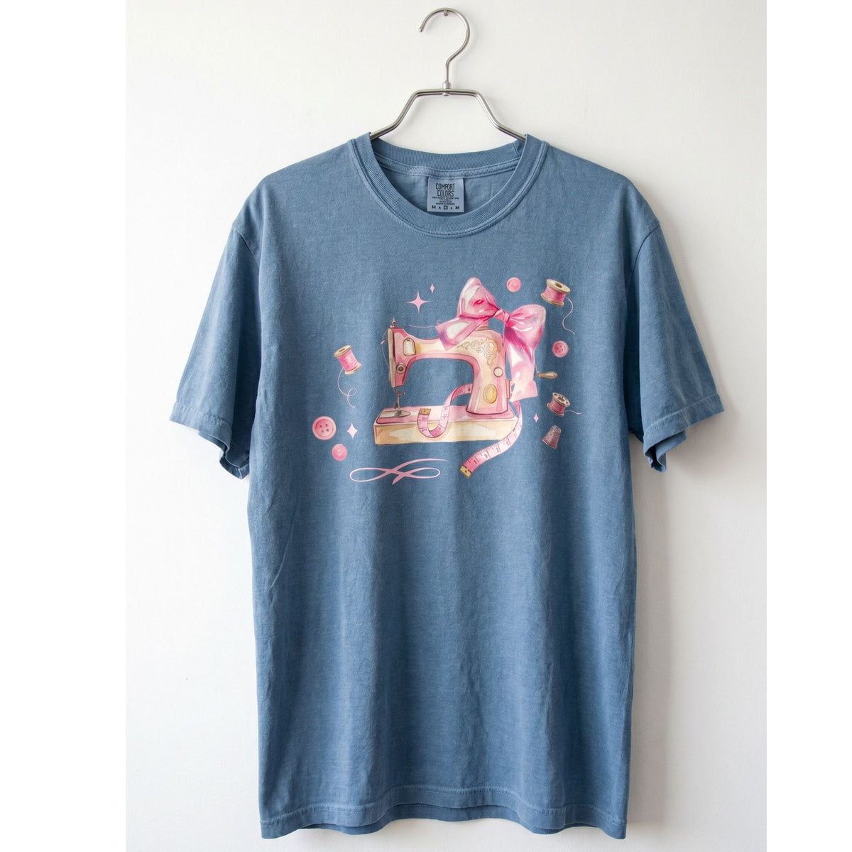 Coquette Sewing T-Shirt in Blue Jean hanging front-facing on a metal hanger against a plain white wall.