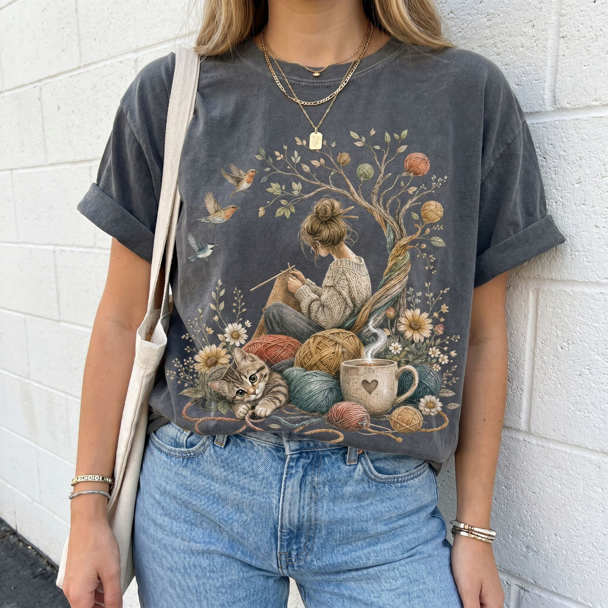 Cottagecore Knitting T-Shirt in Pepper, worn front-facing by a person against a white brick wall with tote bag.
