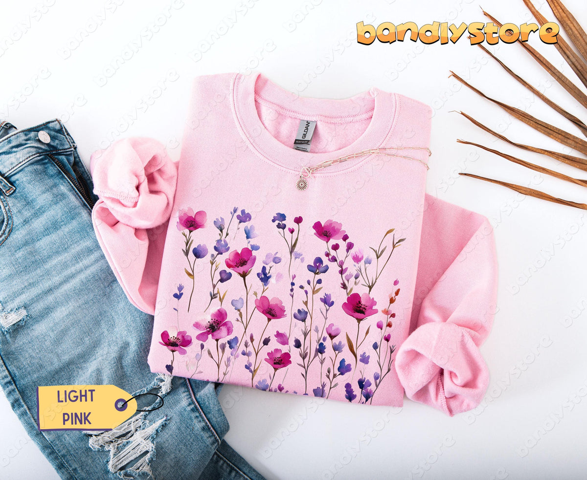 Pressed Wildflowers Sweatshirt in light pink folded flat beside ripped jeans and dried palm leaves.