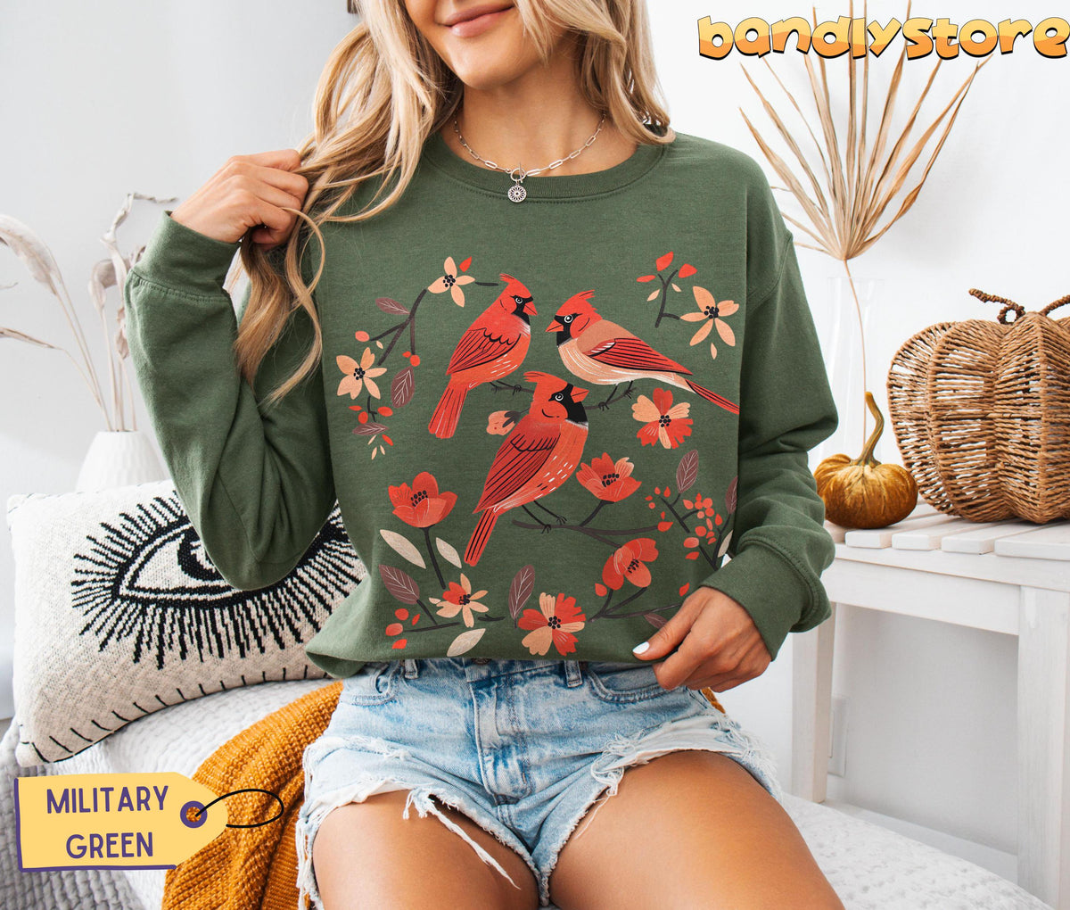 Cottagecore Red Cardinal Sweatshirt in military green on a seated model amid indoor autumn decor and pillows.