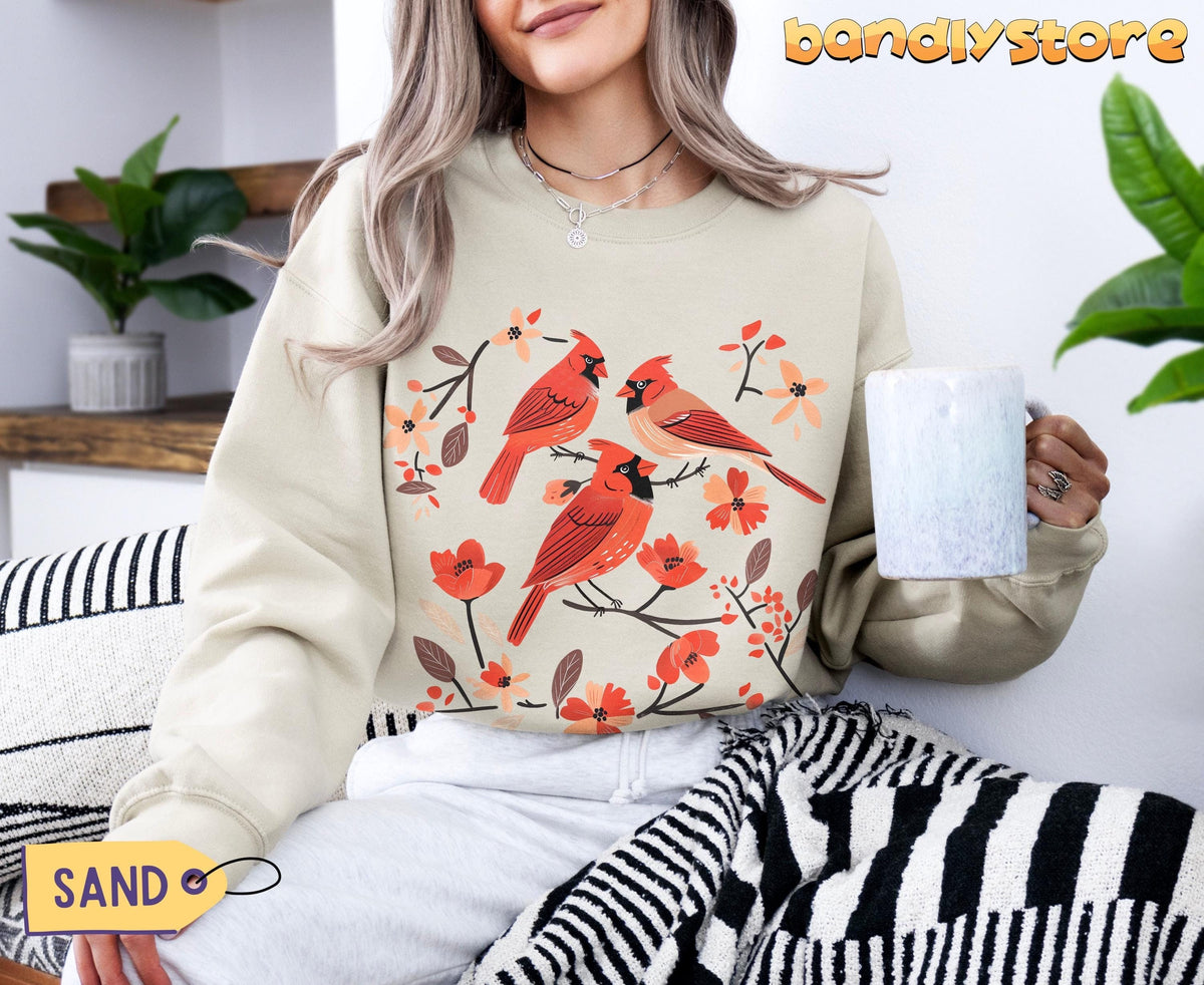 Cottagecore Red Cardinal Sweatshirt in sand worn by a seated person holding a mug beside plants and a striped blanket.