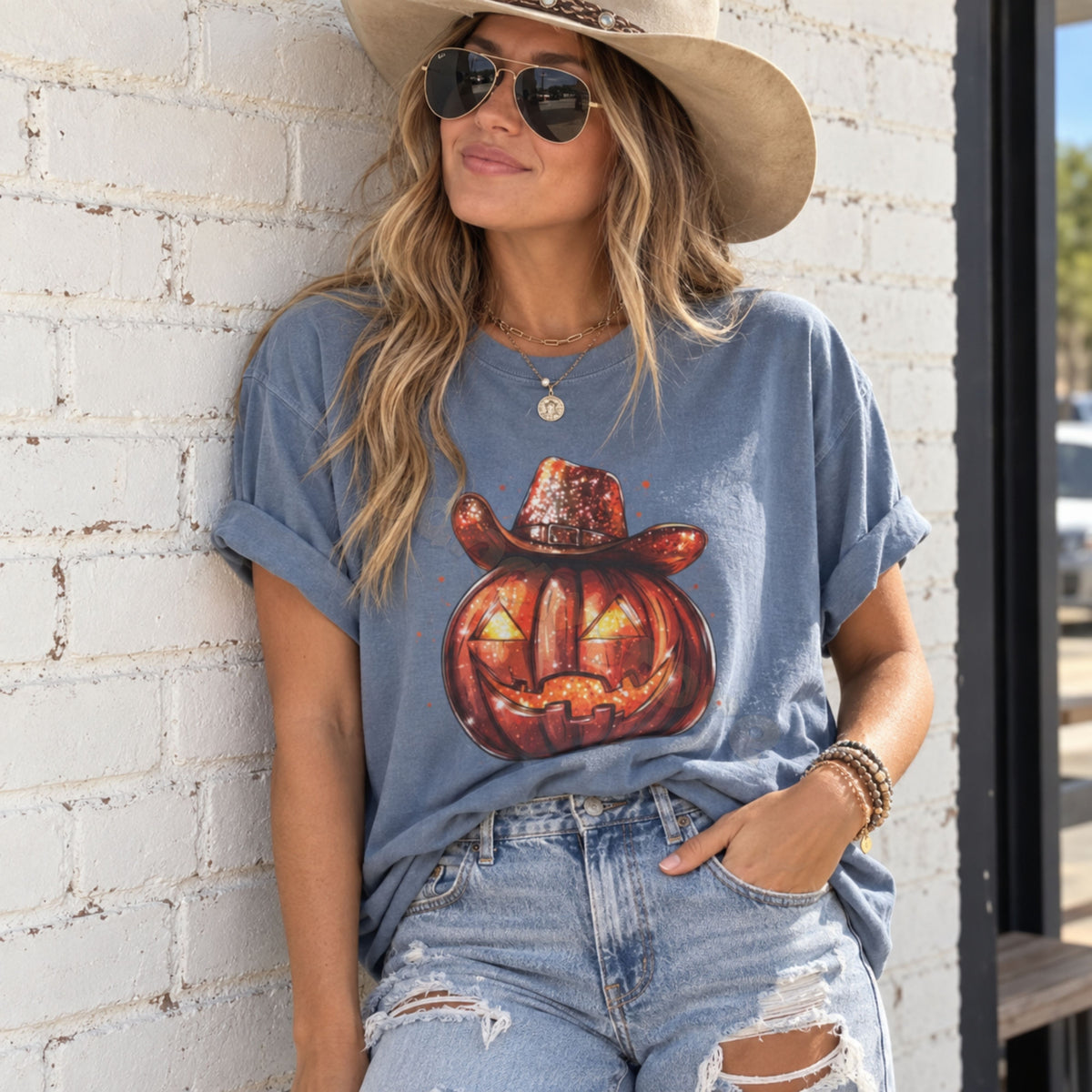 Cowboy Jack-O-Lantern T-Shirt in Blue Jean on woman leaning against white brick wall outdoors.