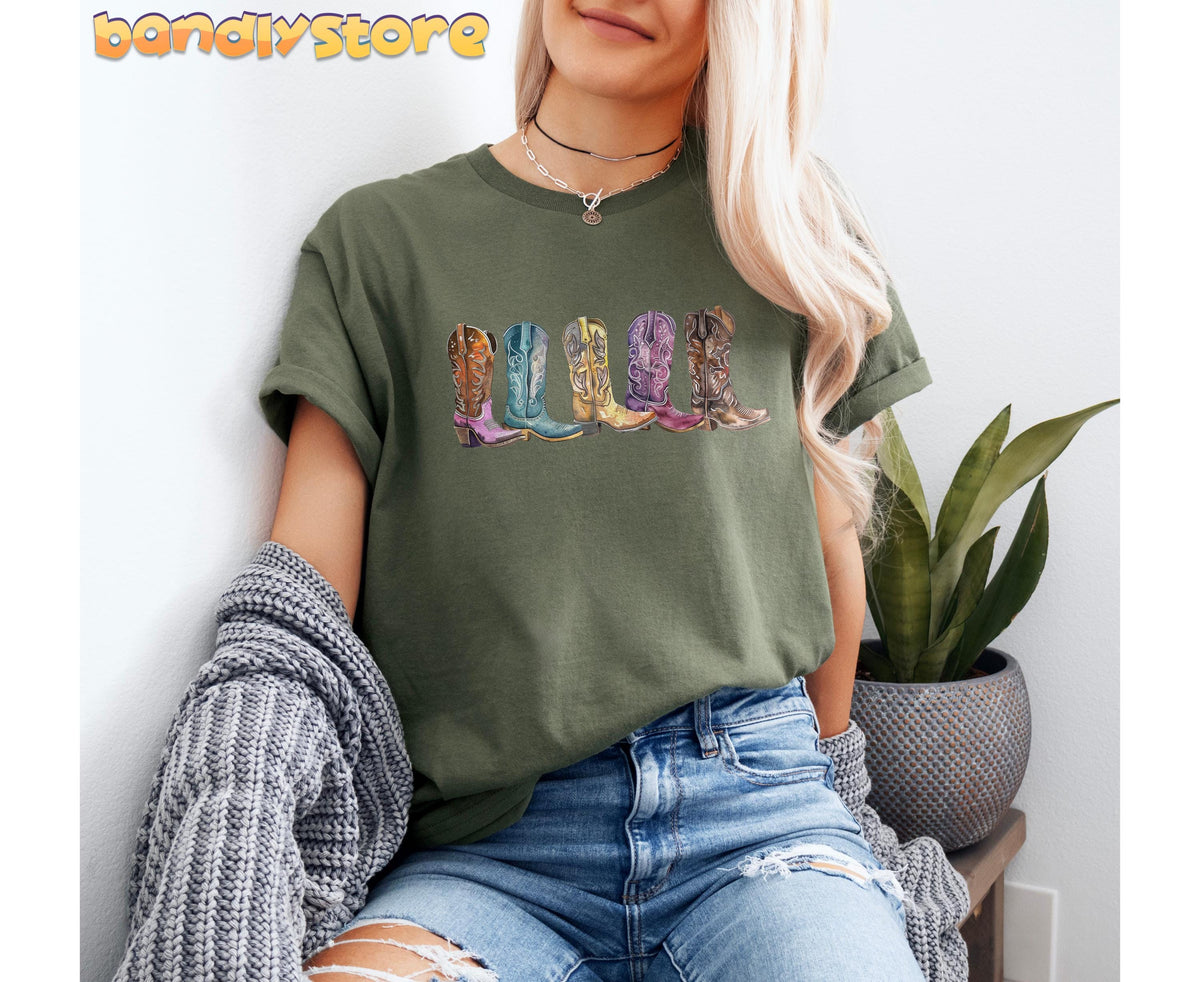Cowgirl Boots T-Shirt in Military Green on a seated model, five colorful boots printed across the chest beside a plant.