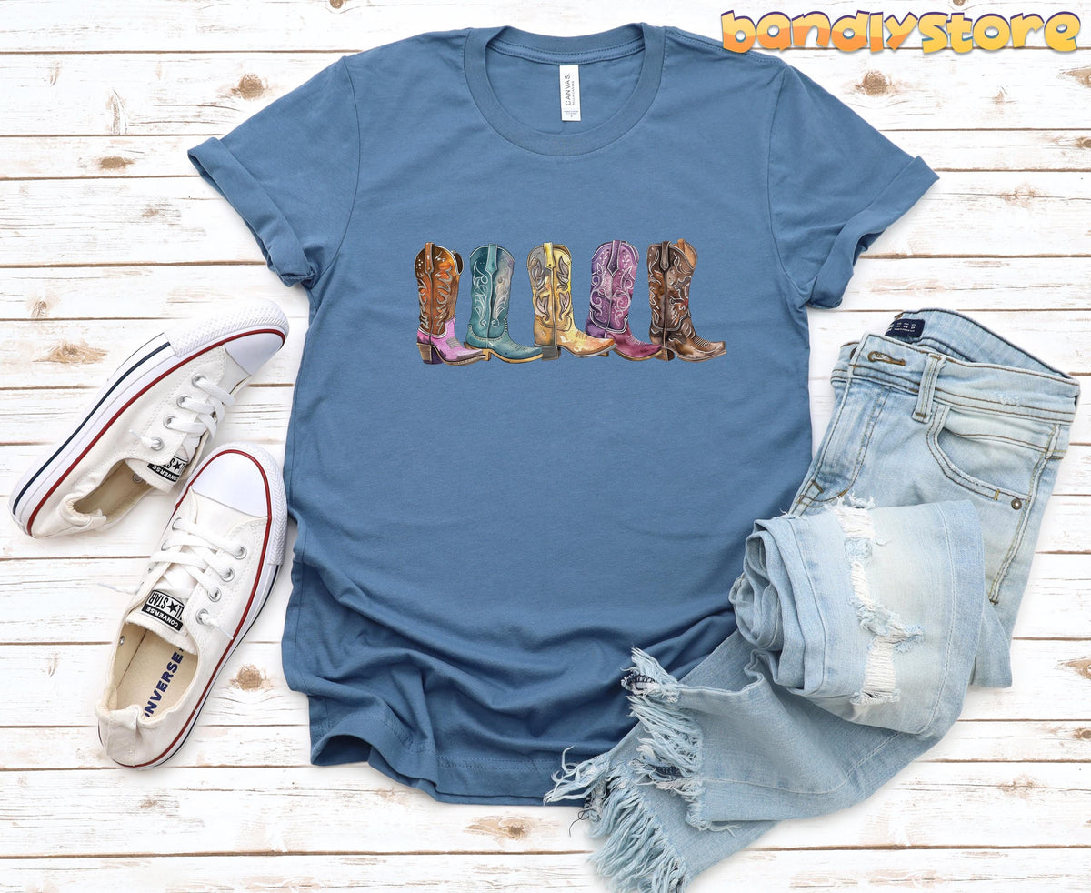 Cowgirl Boots T-Shirt in steel blue flat on wood beside white sneakers and ripped jeans.