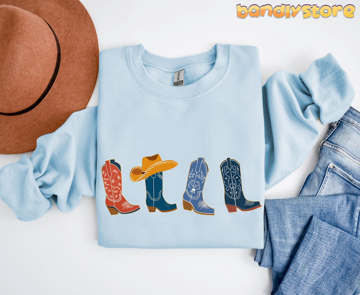 Cowgirl Boots Sweatshirt light blue folded flat with cowboy boots print, brown hat left and blue jeans right.