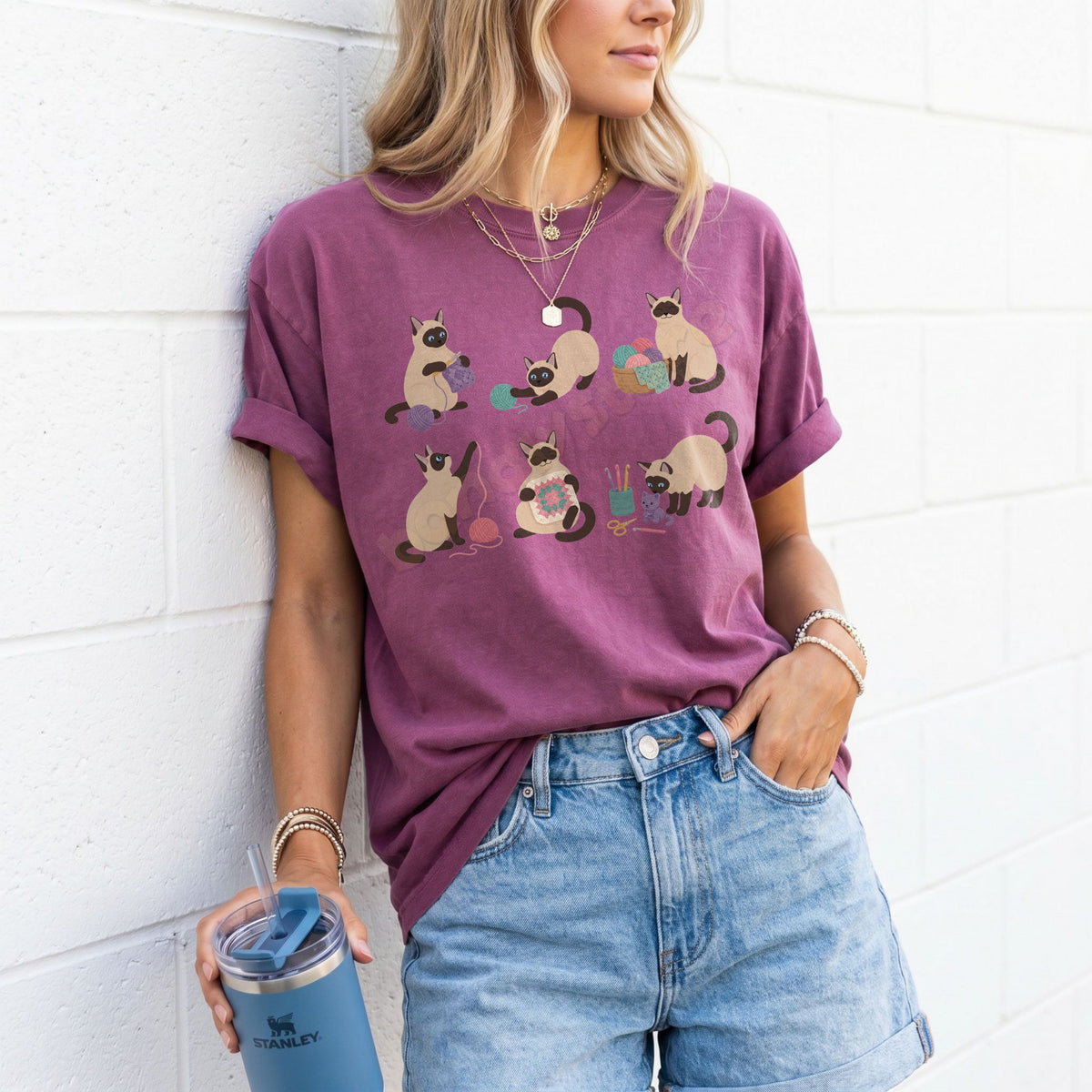 Funny Crocheting Siamese Cats T-Shirt in Berry, worn by a woman leaning on a white brick wall holding a blue tumbler.
