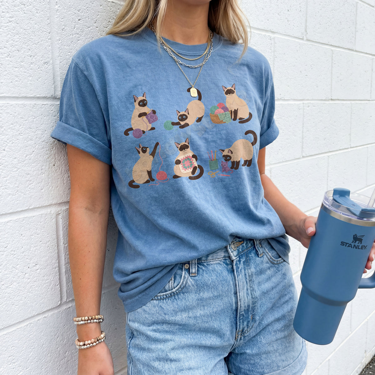 Funny Crocheting Siamese Cats T-Shirt in Blue Jean, worn by a person against a white brick wall holding a blue tumbler.
