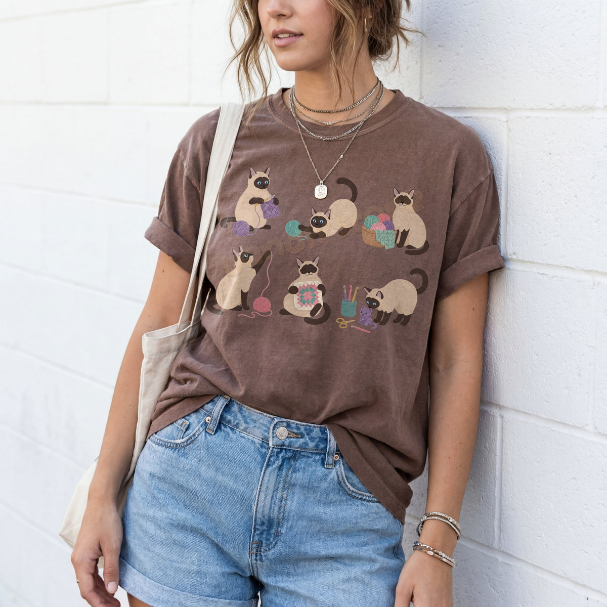 Funny Crocheting Siamese Cats T-Shirt in espresso on a person leaning against a white brick wall with a tote bag.