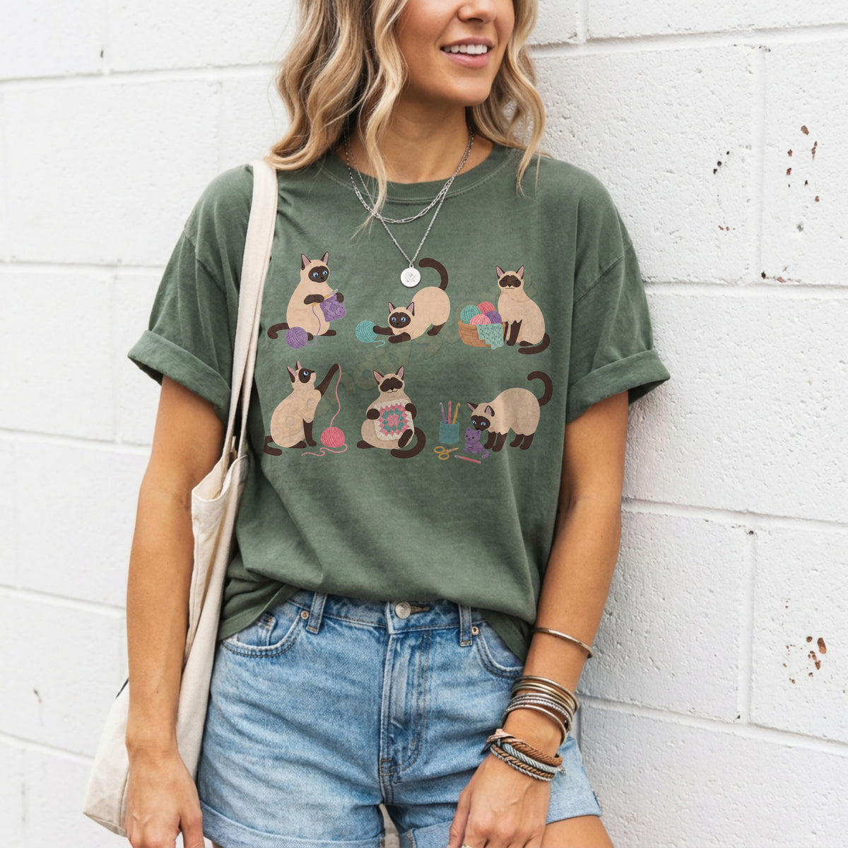 Funny Crocheting Siamese Cats T-Shirt in moss worn by a woman leaning on a white brick wall with tote bag and denim shorts.