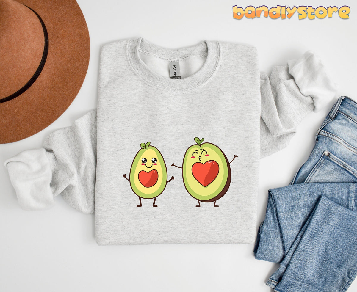 Cute Avocado Love Sweatshirt in ash laid flat beside a brown hat and blue jeans on a white surface.