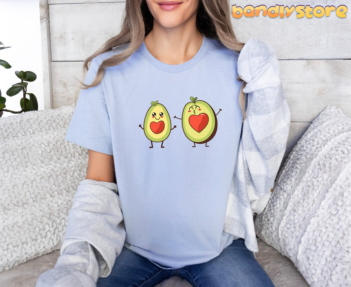 Cute Avocado Love T-Shirt in light blue on a front-facing seated person with pillows, plant and wood wall.