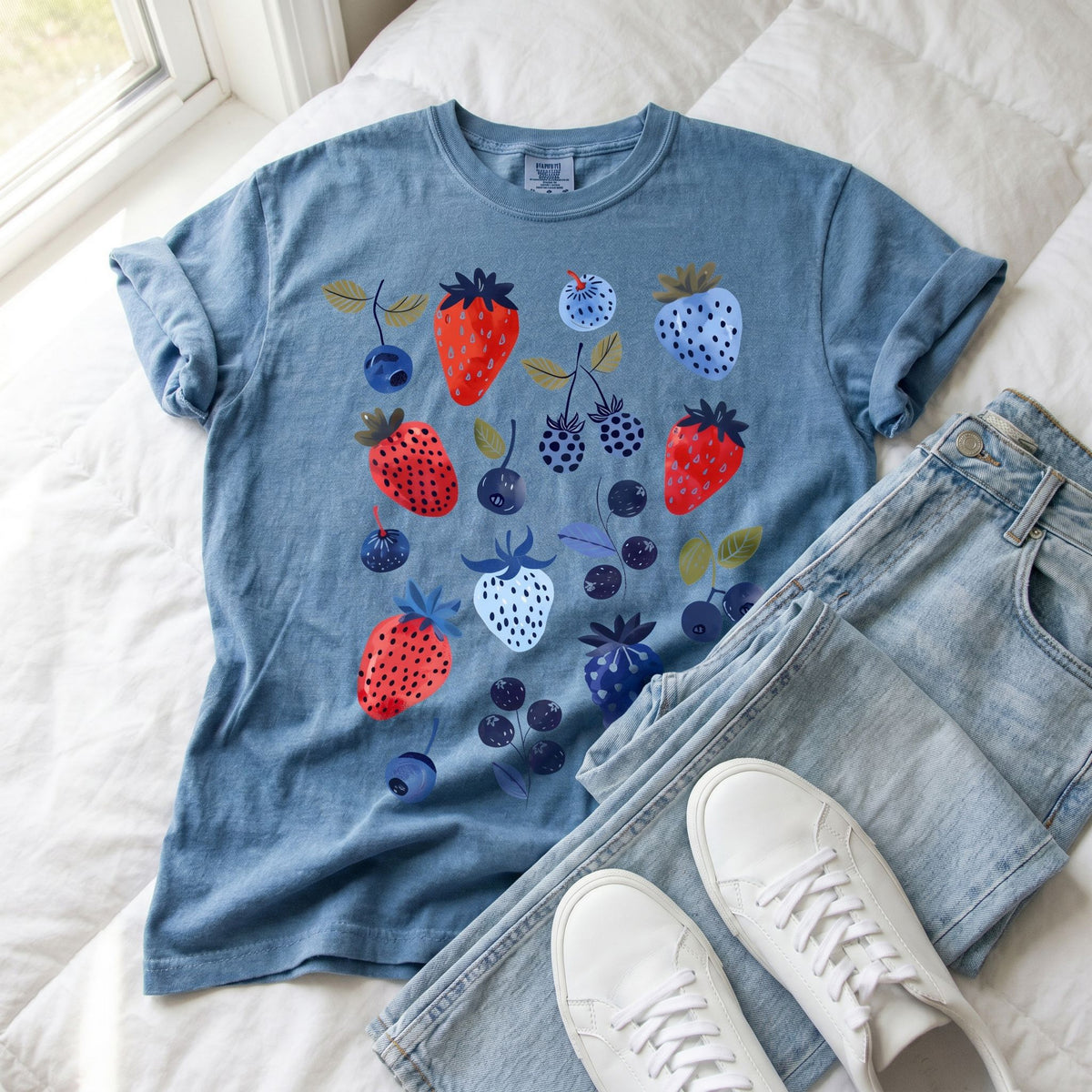 Cute Berry T-Shirt in Blue Jean laid flat on white bedding beside jeans and white sneakers.