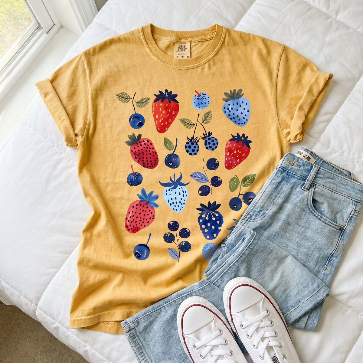 Cute Berry T-Shirt in mustard laid flat on white bedding beside jeans and sneakers.