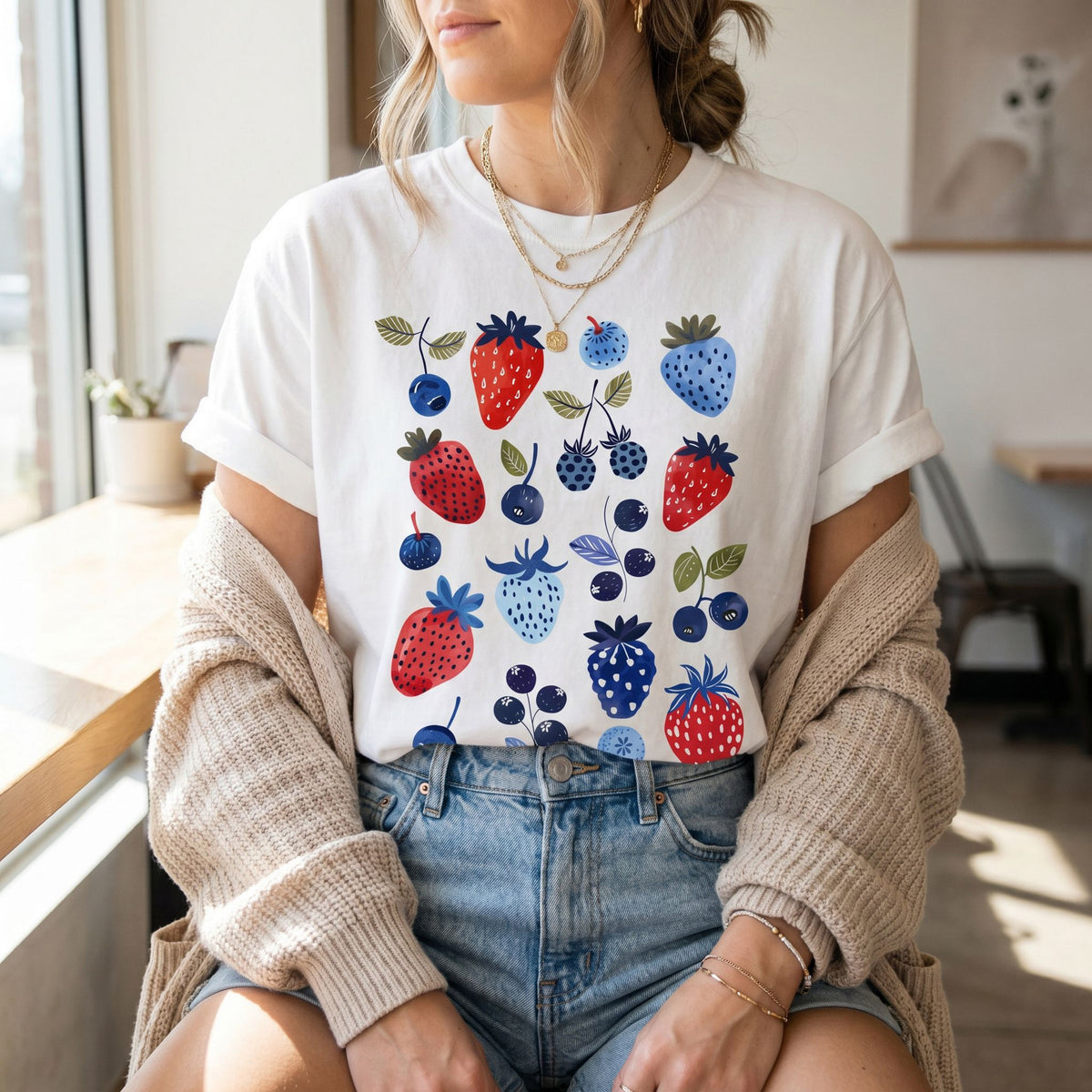 Cute Berry T-Shirt in white, worn by a seated person with gold necklaces, jeans and beige cardigan by a sunny window.